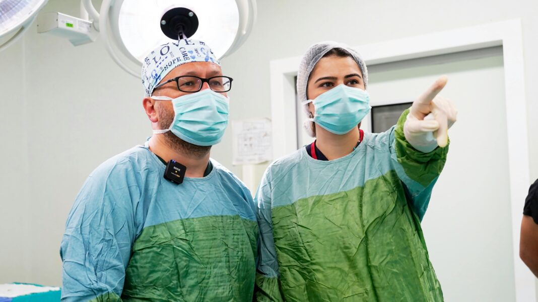 Neuralink’s ‘Blindsight’ to Commence Human Trials: A New Frontier in Cognitive Science Two surgeons in green scrubs and masks discussing a procedure in a modern operating theatre.